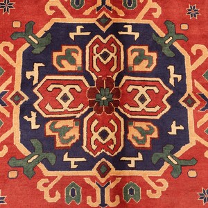 May include: A detailed shot of a patterned rug in red, navy blue, and beige. The central design is a medallion with geometric shapes in red, beige, and green, on a navy blue field. The border is red with repeating floral patterns.