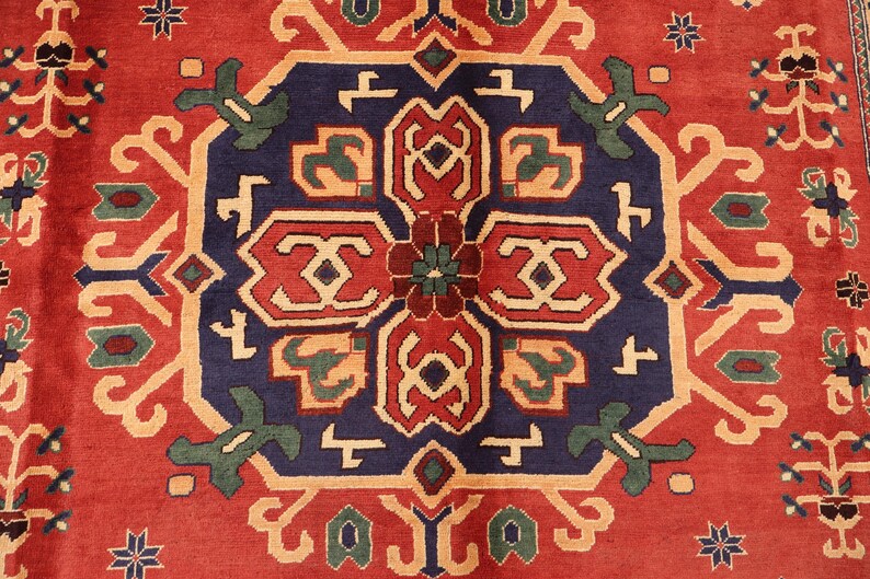 May include: A detailed shot of a patterned rug in red, navy blue, and beige. The central design is a medallion with geometric shapes in red, beige, and green, on a navy blue field. The border is red with repeating floral patterns.