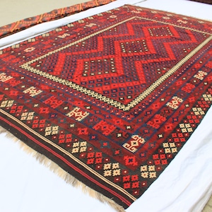 May include: A rectangular, hand-woven rug with a geometric design. The rug features a central diamond pattern in shades of red and burgundy, bordered by intricate patterns in cream, black, and blue. The rug has a fringed edge.