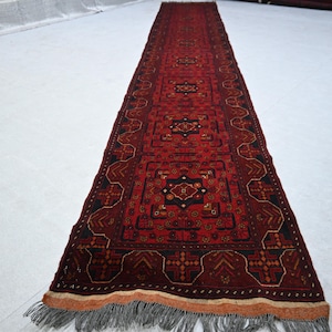Puede incluir: Una alfombra larga y rectangular con un fondo rojo intenso y intrincados patrones geométricos en negro, marrón y dorado. La alfombra tiene un borde con flecos y probablemente está hecha de lana. El diseño es simétrico y tradicional.