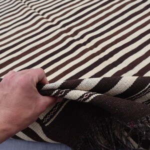 May include: A brown and white striped woven blanket with fringe. The blanket is folded in half and a hand is holding the edge of the blanket.