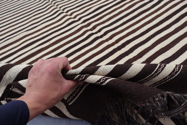 May include: A brown and white striped woven blanket with fringe. The blanket is folded in half and a hand is holding the edge of the blanket.
