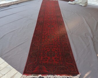Könnte beinhalten: Ein langer, rechteckiger roter Teppich mit einem komplizierten geometrischen Muster in Dunkelrot und Braun. Der Teppich hat einen Fransen an den kurzen Enden und wird auf einer grauen Oberfläche gezeigt. Der Teppich ist im traditionellen Stil.