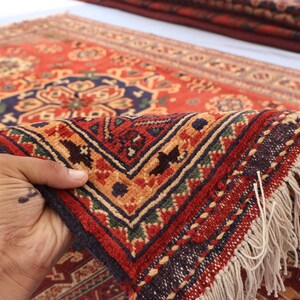 May include: A close-up of a red, patterned rug with a detailed geometric design. The rug features a dark blue border, tan fringe, and a complex pattern in shades of red, orange, and green. The rug is being held up, showcasing its texture.