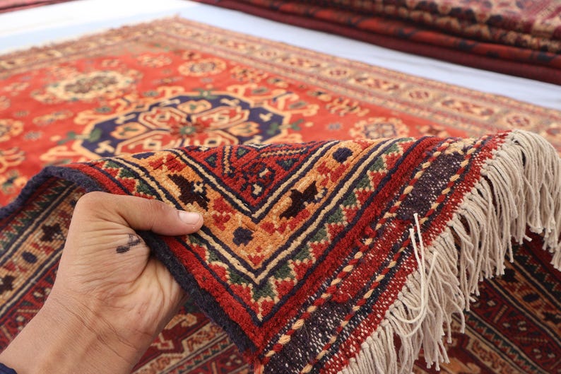 May include: A close-up of a red, patterned rug with a detailed geometric design. The rug features a dark blue border, tan fringe, and a complex pattern in shades of red, orange, and green. The rug is being held up, showcasing its texture.