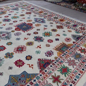 May include: A large, off-white rug with a repeating pattern of colorful geometric shapes. The design includes squares, stars, and floral motifs in shades of red, blue, green, and yellow. A detailed border frames the rug.