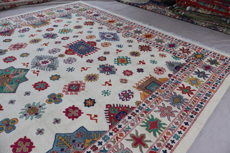 May include: A large, off-white rug with a repeating pattern of colorful geometric shapes. The design includes squares, stars, and floral motifs in shades of red, blue, green, and yellow. A detailed border frames the rug.