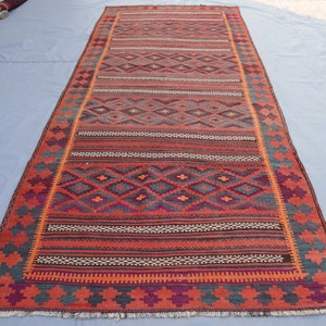 May include: A traditional handwoven rug with a geometric pattern in shades of red, orange, blue, and purple. The rug has a fringed edge and is made of wool.