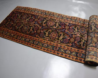 Puede incluir: Una alfombra rectangular tipo pasillo con fondo negro y un intrincado patrón de flores y enredaderas en tonos rojo oscuro, azul y dorado. La alfombra está parcialmente enrollada en un extremo, mostrando un borde detallado con motivos.