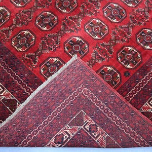 May include: A red oriental rug with a geometric pattern. The rug features a central field with repeating octagonal motifs in shades of red, brown, and cream. The border is a darker red with intricate designs. The rug is folded to show the underside.