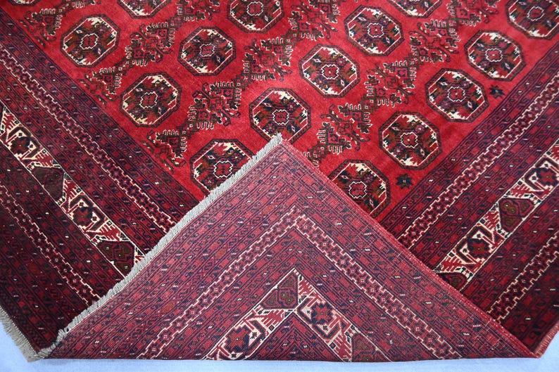 May include: A red oriental rug with a geometric pattern. The rug features a central field with repeating octagonal motifs in shades of red, brown, and cream. The border is a darker red with intricate designs. The rug is folded to show the underside.