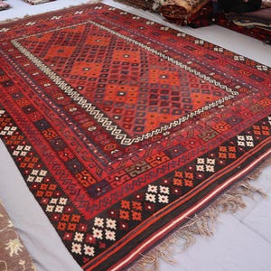 May include: A large, rectangular red rug with an intricate geometric pattern. The rug features a central diamond design in shades of red and orange, framed by borders with repeating patterns and fringe edges. The rug measures approximately 9 x 12 feet.