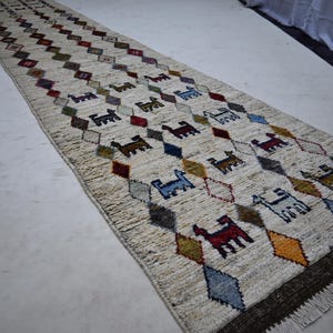 May include: A long, narrow runner rug featuring a cream-coloured base with a repeating diamond pattern. Inside the diamonds are small, stylised animal figures in various colours including red, blue, brown, and yellow. The rug has a dark brown fringe.