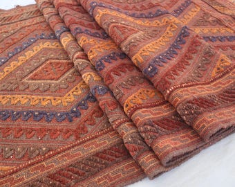 Könnte beinhalten: Gefalteter Teppich mit detailliertem geometrischem Muster. Der Teppich zeigt ein Rautenmuster in Orange-, Rot-, Braun- und Blautönen. Die Stickerei und Textur sind deutlich sichtbar.