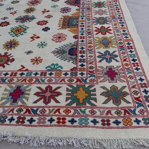 May include: A large, off-white rug with a detailed pattern. The rug features a repeating design of colorful geometric shapes in red, blue, green, yellow, and pink. A fringed edge completes the design.