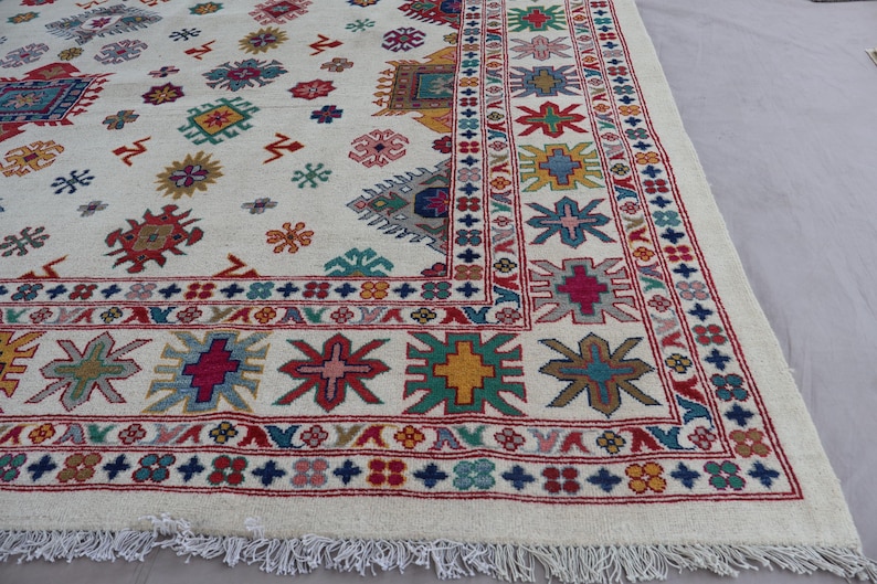 May include: A large, off-white rug with a detailed pattern. The rug features a repeating design of colorful geometric shapes in red, blue, green, yellow, and pink. A fringed edge completes the design.