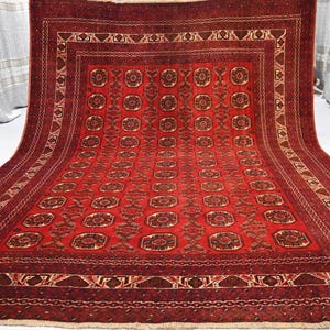 May include: A rectangular red rug with an intricate geometric pattern. The rug features a central field of repeating medallions and a detailed border with various shapes and designs. The color palette includes shades of red, brown, and cream.