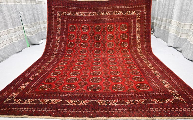 May include: A rectangular red rug with an intricate geometric pattern. The rug features a central field of repeating medallions and a detailed border with various shapes and designs. The color palette includes shades of red, brown, and cream.