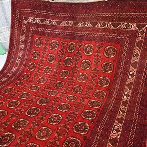 May include: A red and brown patterned rug with a repeating geometric design. The rug features a central field of repeating medallions and a detailed border. The rug is made of a woven material and has a rich, textured appearance.