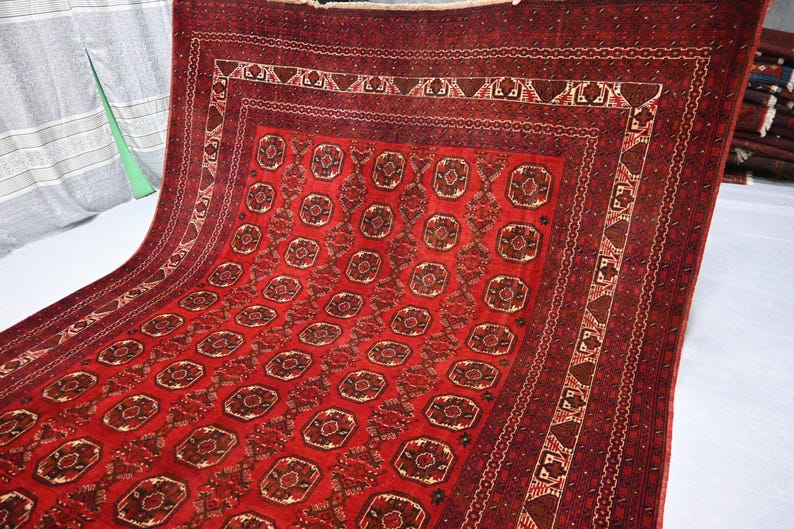 May include: A red and brown patterned rug with a repeating geometric design. The rug features a central field of repeating medallions and a detailed border. The rug is made of a woven material and has a rich, textured appearance.