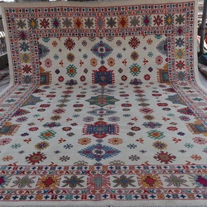 May include: A large, rectangular rug with a cream-colored background and a repeating pattern of colorful geometric shapes. The border features a detailed design in red, blue, and yellow. The rug is displayed outdoors.