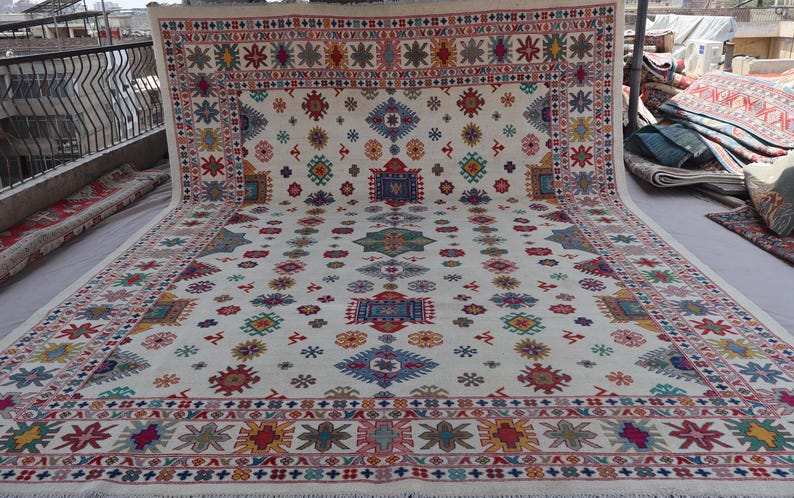 May include: A large, rectangular rug with a cream-colored background and a repeating pattern of colorful geometric shapes. The border features a detailed design in red, blue, and yellow. The rug is displayed outdoors.