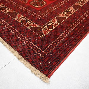 May include: Close-up of a red and beige patterned rug. The rug features a geometric design with intricate details in shades of red, brown, and beige. The edges are finished with a fringe.