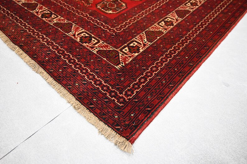 May include: Close-up of a red and beige patterned rug. The rug features a geometric design with intricate details in shades of red, brown, and beige. The edges are finished with a fringe.