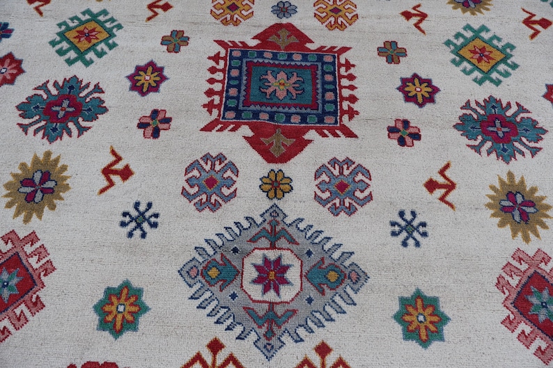 May include: A cream-colored rug with a repeating pattern of colorful geometric shapes. The design includes squares, diamonds, and floral motifs in shades of red, blue, yellow, and green. The rug has a textured surface.