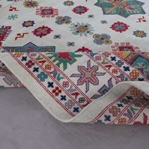 May include: A rolled-up, off-white rug with a detailed geometric pattern. The design features various shapes in red, blue, yellow, and green, with a red border. The rug's intricate design adds a touch of elegance.