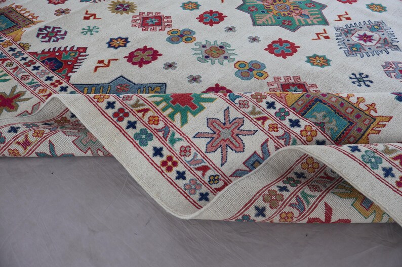 May include: A rolled-up, off-white rug with a detailed geometric pattern. The design features various shapes in red, blue, yellow, and green, with a red border. The rug's intricate design adds a touch of elegance.