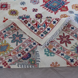 May include: Cream-colored rug with a geometric pattern in red, blue, yellow, and green. The rug has a detailed border and fringe. The design features repeating floral and geometric shapes.