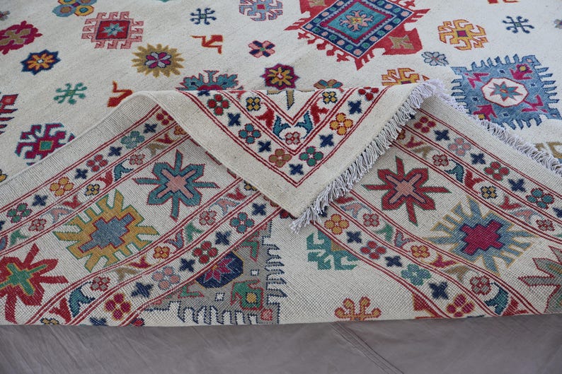 May include: Cream-colored rug with a geometric pattern in red, blue, yellow, and green. The rug has a detailed border and fringe. The design features repeating floral and geometric shapes.