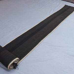 May include: A long, rolled-up black runner rug with a central dark stripe and cream-colored trim. The rug is partially unrolled, revealing its texture and design. The rug is on a light gray surface.