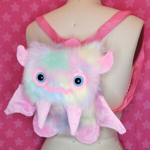 Może przedstawiać: Pluszowy plecak w pastelowych kolorach tęczy w kształcie potwora z dużymi oczami, r&oacute;żowymi rogami i skrzydłami.