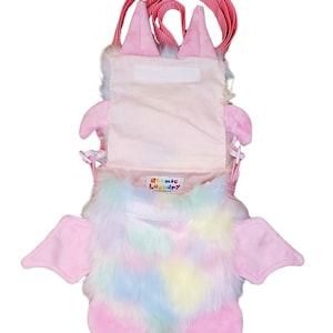 Może przedstawiać: R&oacute;żowa pluszowa torebka na ramię w kształcie nietoperza z tęczowym futerkiem i r&oacute;żowym paskiem. Torebka ma zamek błyskawiczny i małą etykietę z napisem "Atomic Laundry".