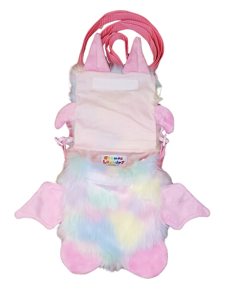 Może przedstawiać: R&oacute;żowa pluszowa torebka na ramię w kształcie nietoperza z tęczowym futerkiem i r&oacute;żowym paskiem. Torebka ma zamek błyskawiczny i małą etykietę z napisem "Atomic Laundry".