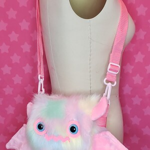 Może przedstawiać: R&oacute;żowa i tęczowa, pluszowa torebka na ramię w kształcie nietoperza z dużymi oczami i zębami. Torebka ma regulowany r&oacute;żowy pasek.