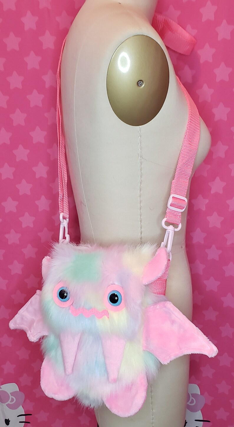 Może przedstawiać: R&oacute;żowa i tęczowa, pluszowa torebka na ramię w kształcie nietoperza z dużymi oczami i zębami. Torebka ma regulowany r&oacute;żowy pasek.