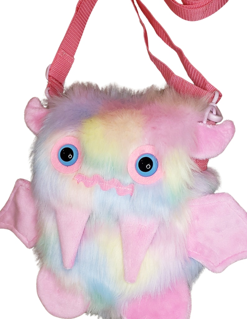 Może przedstawiać: R&oacute;żowa i tęczowa pluszowa torba na ramię w kształcie potwora z dużymi niebieskimi oczami i r&oacute;żowym paskiem.