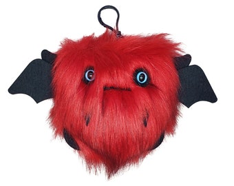 May include: A plush, red monster with black felt wings and horns. The monster has large blue eyes and a small, stitched mouth. A black loop is attached to the top for hanging. The toy is made of soft, furry material.