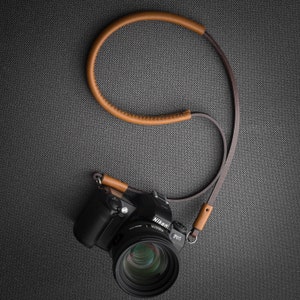 May include: A black Nikon D780 camera with a brown leather camera strap. The camera has a lens attached and the strap is looped around the camera and hanging down.