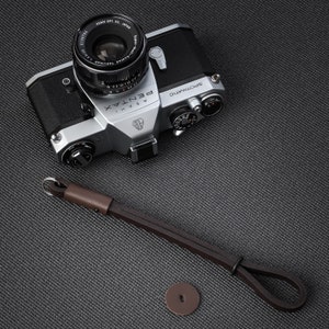 Può includere: Una macchina fotografica a pellicola da 35 mm di colore argento e nero con un cinturino in pelle nera. La macchina fotografica ha un obiettivo argentato con il testo "ASAHI PENTAX" e "SPOTMATIC" stampato su di esso.