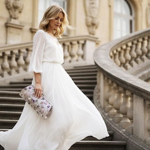May include: A flowing white dress with long sleeves and a cinched waist. The dress is paired with a floral clutch. The woman is descending a stone staircase with a curved railing, creating a classic and elegant look.