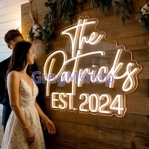 Puede incluir: Un letrero de neón con las palabras "The Patricks EST. 2024" en una fuente cursiva. El letrero está iluminado con una luz blanca cálida y montado en una pared de madera. Una pareja está cerca.