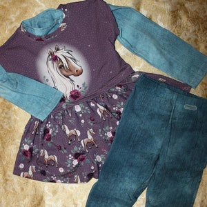 May include: A children's outfit comprising a purple top with a horse design and a floral skirt, paired with denim-style leggings. The top has light blue sleeves. The outfit is displayed on a gold patterned surface. Measurements are in cm.