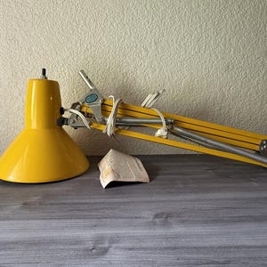 May include: A yellow articulated desk lamp with a conical shade and a folding metal arm mechanism. The lamp features a white power cord wrapped around the frame, silver-toned metal springs, and a small paper document placed near the base.
