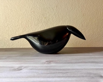 May include: A smooth, solid black bird figurine with a minimalist, rounded body shape, a pointed beak, and a tapered tail. The surface of the object is highly reflective, showing subtle highlights and a glossy finish throughout the entire form.
