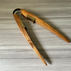 May include: A wooden nutcracker with brass accents. The nutcracker has a curved handle and two long, pointed arms. The brass pieces are serrated. The wood has a natural grain and a warm, brown color.