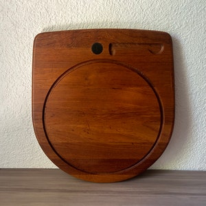 May include: A wooden cutting board with a unique design. The board is a warm brown color and features a circular indentation in the center. It has a small circular indentation and a groove for a knife. The board measures approximately 12 inches wide.